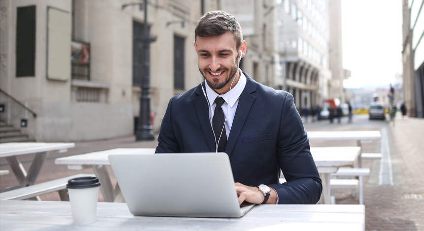 A businessman with earphones in sitting at a table at a cafe outside looking at his laptop and smiling.
